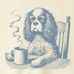 Pode incluir: Camiseta creme com uma ilustração azul de um Cavalier King Charles Spaniel. O cão está sentado em uma mesa com uma caneca de café, com uma expressão cansada. A camiseta tem gola redonda e mangas curtas.