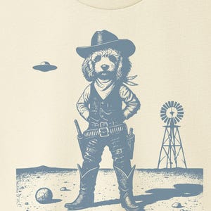 Puede incluir: Camiseta color crema con un gráfico azul de un perro vestido de vaquero, con sombrero, chaleco y botas. También se representa un OVNI y un molino de viento. La camiseta tiene cuello redondo y mangas cortas. El logo de la marca "TACO DOG" está en la parte superior derecha.