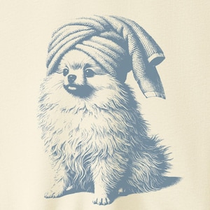 May include: Cream-colored t-shirt featuring a blue illustration of a Pomeranian dog wearing a towel on its head. The dog is sitting, with fluffy fur and a playful expression. The shirt is hanging on a wooden hanger.