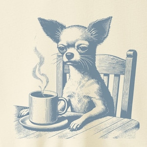 May include: Cream-colored t-shirt featuring a blue illustration of a Chihuahua sitting at a table, holding a coffee cup. The dog is depicted with a serious expression, and steam rises from the coffee. The shirt has a crew neck and short sleeves.