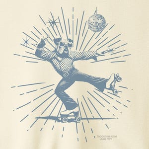 Pode incluir: Camiseta creme com um gráfico azul de um cão em patins, com uma bola de discoteca e um design de explosão estelar. O texto "TACO DOG" está em um círculo no canto superior direito. O texto "TACODOG.COM JUNE 1979" está na parte inferior.