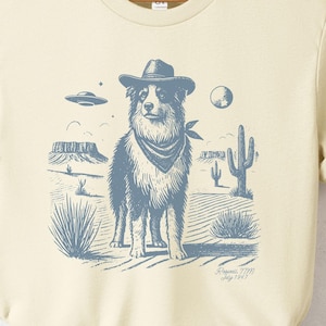 May include: A cream-coloured t-shirt featuring a blue illustration of a dog wearing a cowboy hat and bandana. The dog is standing in a desert landscape with cacti and a UFO in the background. The text "Space Cowboy" is printed below the illustration.