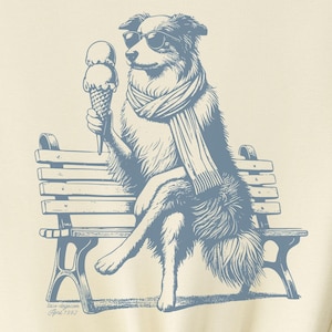 May include: A light yellow t-shirt featuring a blue illustration of a dog wearing sunglasses and a scarf, sitting on a park bench and holding an ice cream cone. The text "Taco Dog" is in the top right corner of the image.