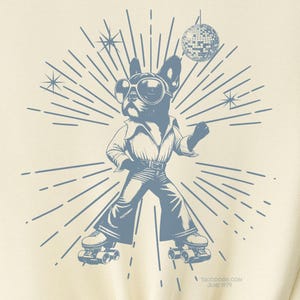 May include: A light yellow t-shirt with a blue graphic of a dog wearing roller skates and holding a disco ball. The dog is wearing sunglasses and a disco outfit. The graphic is surrounded by a sunburst design.