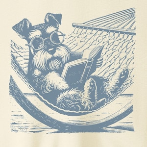 K&ouml;nnte beinhalten: Ein cremefarbenes T-Shirt mit einer blauen Illustration eines Hundes, der eine Brille tr&auml;gt und ein Buch liest, w&auml;hrend er in einer H&auml;ngematte entspannt.