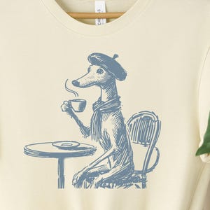 May include: A light beige t-shirt featuring a blue illustration of a dog wearing a beret and holding a cup of coffee. The dog is seated at a table with a plate and a chair. The text "Taco Dog" is in the top right corner of the image.