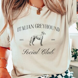 以下が含まれることがあります： 「ITALIAN GREYHOUND EST. 1886 Social Club」と黒で書かれたグラフィックデザインが施されたベージュのTシャツ。グラフィックには、イタリアングレイハウンドの白黒のイラストも含まれています。