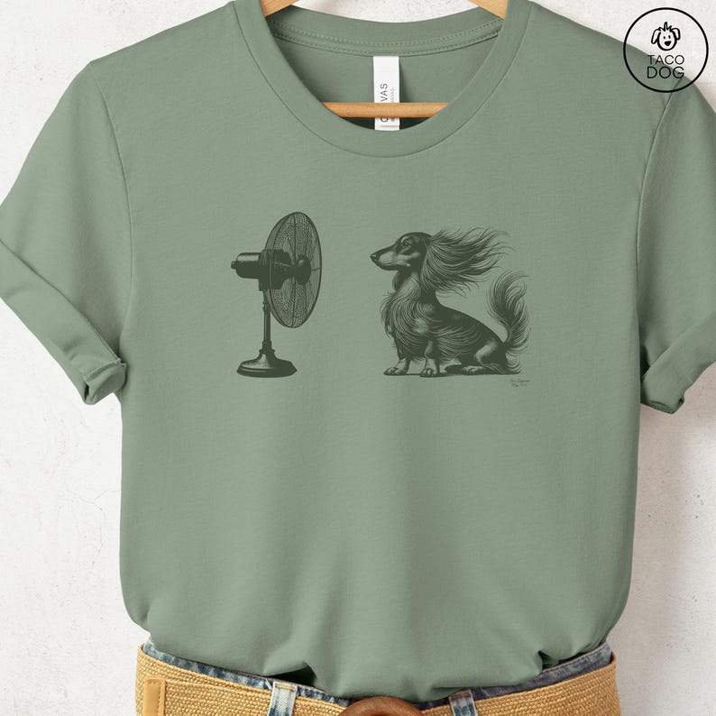 May include: Sage green t-shirt featuring a black and white illustration of a long-haired dachshund with wind-blown fur, positioned near a standing fan. The shirt has a crew neck and short sleeves. A Taco Dog logo is visible on the upper right.