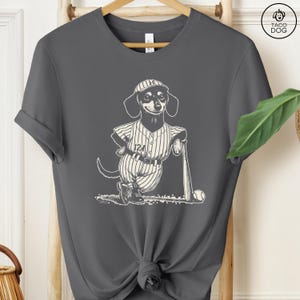 May include: A gray t-shirt with a black and white illustration of a dachshund wearing a baseball uniform and holding a bat. The dachshund is wearing a baseball cap with the letter "H" on it. The text "124" is on the uniform.