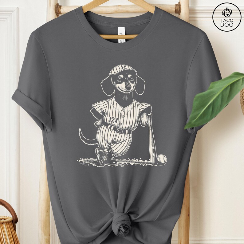 May include: A gray t-shirt with a black and white illustration of a dachshund wearing a baseball uniform and holding a bat. The dachshund is wearing a baseball cap with the letter "H" on it. The text "124" is on the uniform.