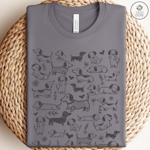 May include: A gray t-shirt with a repeating pattern of black dachshund dogs in various poses. The shirt has a crew neck and short sleeves. The text "Taco Dog" is printed in the top right corner.
