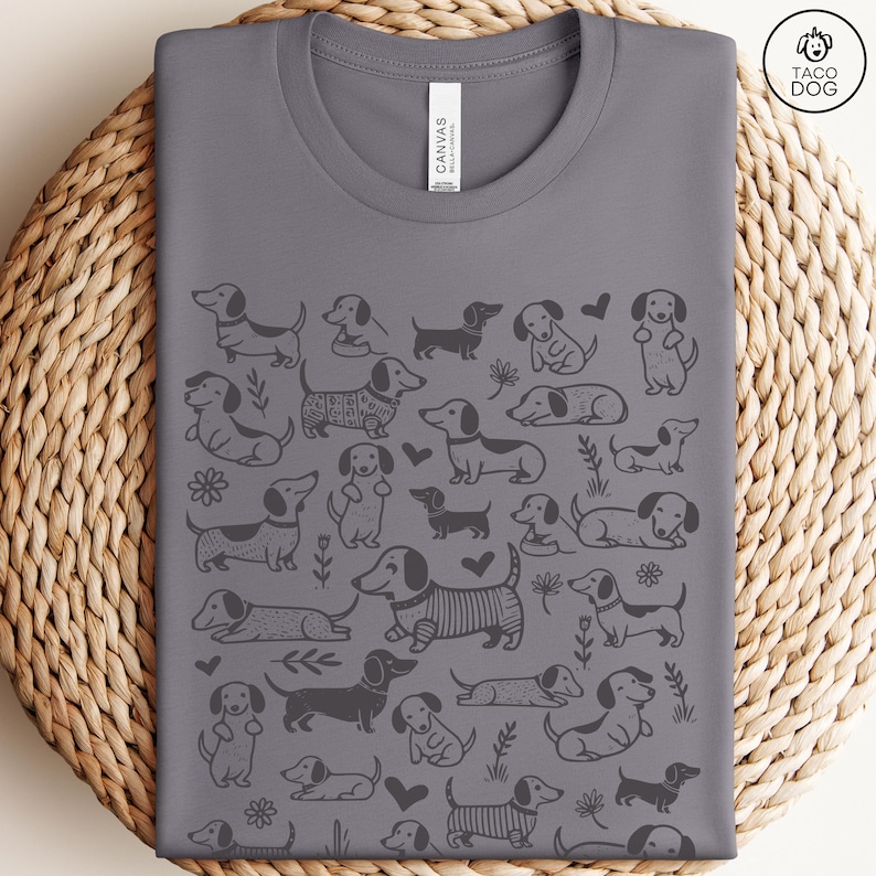 May include: A gray t-shirt with a repeating pattern of black dachshund dogs in various poses. The shirt has a crew neck and short sleeves. The text "Taco Dog" is printed in the top right corner.
