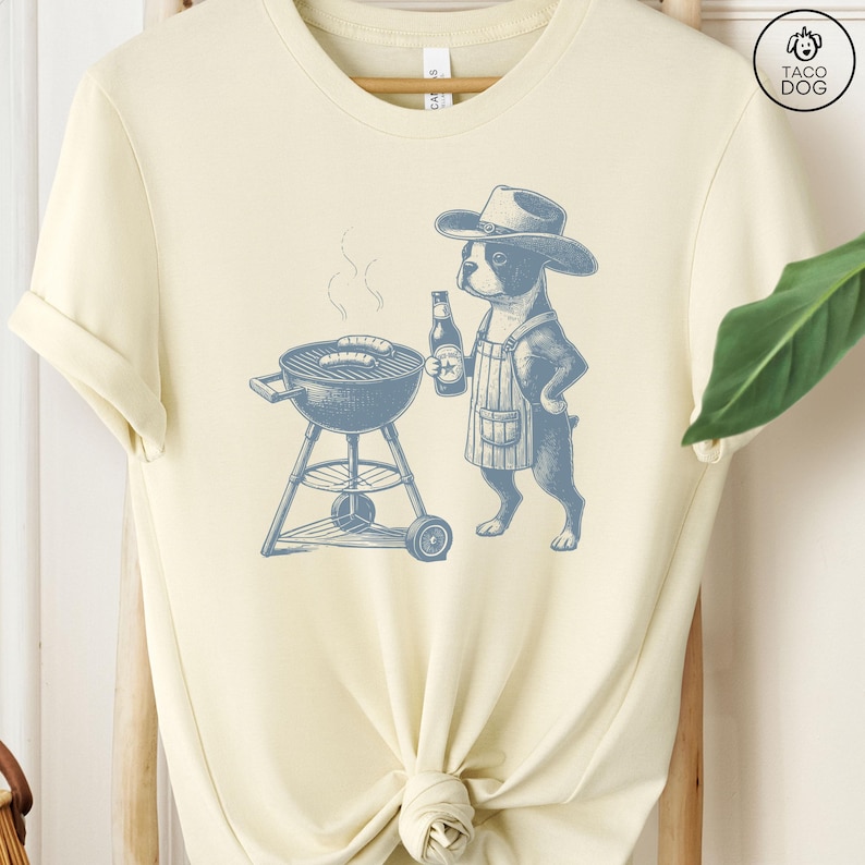 Peut inclure: Un t-shirt jaune clair avec une illustration bleue d'un chien portant un chapeau de cowboy et un tablier, faisant griller des saucisses sur un barbecue au charbon de bois. Le chien tient une bouteille de bi&egrave;re. Le texte "Taco Dog" est dans le coin sup&eacute;rieur droit de l'image.