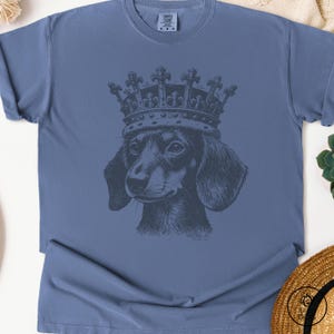May include: A blue t-shirt with a black and white illustration of a dachshund wearing a crown. The crown has a detailed design with ornate details.
