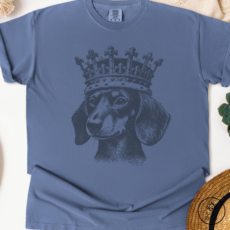 May include: A blue t-shirt with a black and white illustration of a dachshund wearing a crown. The crown has a detailed design with ornate details.