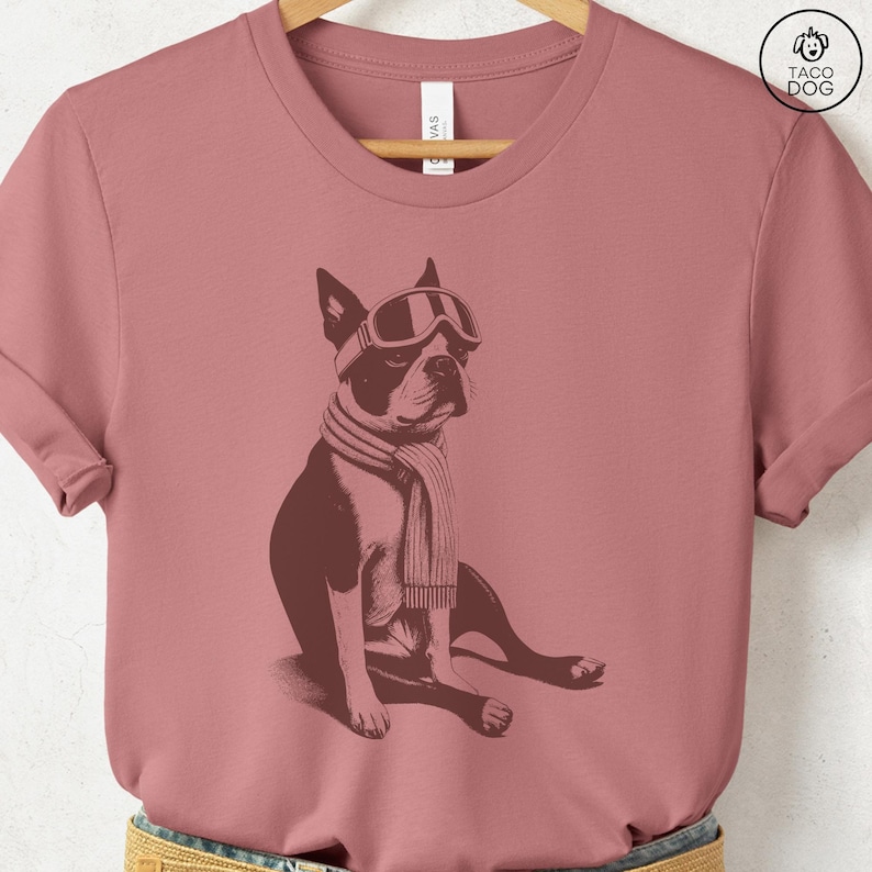 Puede incluir: Camiseta rosa polvoriento con una ilustraci&oacute;n en sepia de un Boston Terrier sentado con gafas de esqu&iacute; y una bufanda. La camiseta tiene cuello redondo y mangas cortas. El logotipo de la marca "TACO DOG" est&aacute; en la esquina superior derecha.