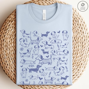 May include: Light blue t-shirt with a pattern of blue dachshund dogs on a white background. The shirt has a crew neck and short sleeves. The text "CANVAS" is visible on the tag.