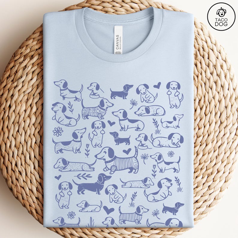 May include: Light blue t-shirt with a pattern of blue dachshund dogs on a white background. The shirt has a crew neck and short sleeves. The text "CANVAS" is visible on the tag.
