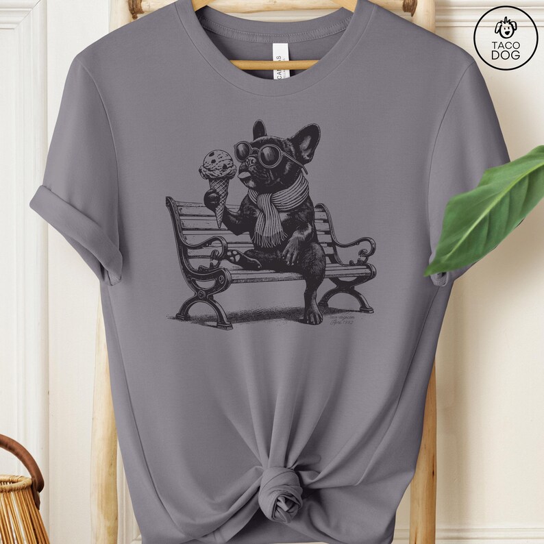 May include: Gray t-shirt featuring a black and white illustration of a French Bulldog wearing sunglasses and a scarf, sitting on a bench and eating an ice cream cone. The shirt has a crew neck and short sleeves. The Taco Dog logo is in the upper right corner.