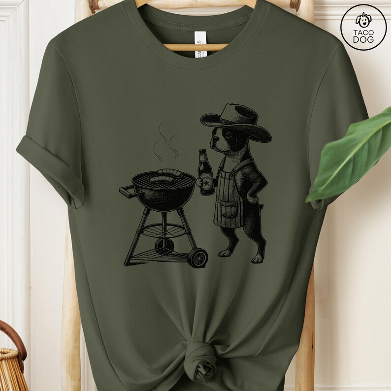 Peut inclure: Un t-shirt vert avec une illustration noire d'un chien portant un chapeau de cowboy et un tablier, grillant des saucisses sur un barbecue au charbon de bois. Le chien tient une bouteille de bi&egrave;re.