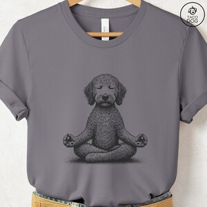 May include: A gray t-shirt featuring a detailed illustration of a dog in a meditative pose. The dog is depicted with closed eyes and crossed legs, hands resting in a yoga mudra. The shirt has a crew neck and short sleeves.