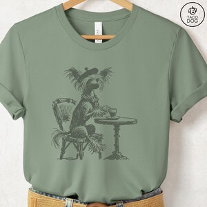 May include: A green t-shirt with a black and white illustration of a dog wearing a beret and sitting at a table with a cup of coffee. The dog is a Chinese Crested.