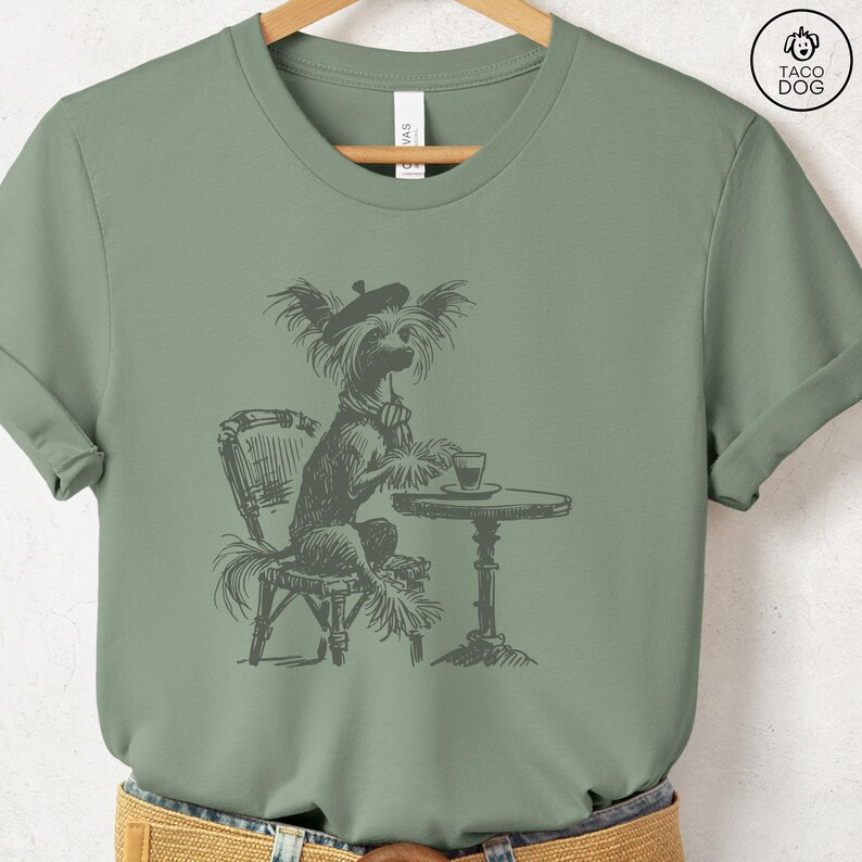 May include: A green t-shirt with a black and white illustration of a dog wearing a beret and sitting at a table with a cup of coffee. The dog is a Chinese Crested.