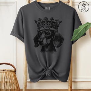 May include: A dark gray t-shirt with a black and white illustration of a dachshund wearing a crown. The shirt has a crew neck and short sleeves.