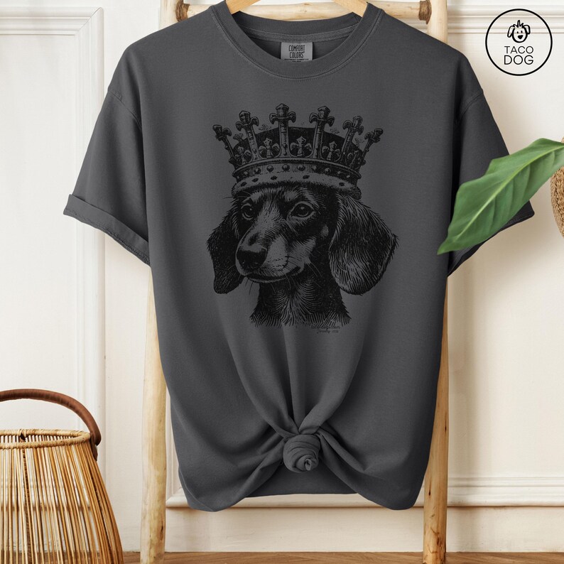 May include: A dark gray t-shirt with a black and white illustration of a dachshund wearing a crown. The shirt has a crew neck and short sleeves.