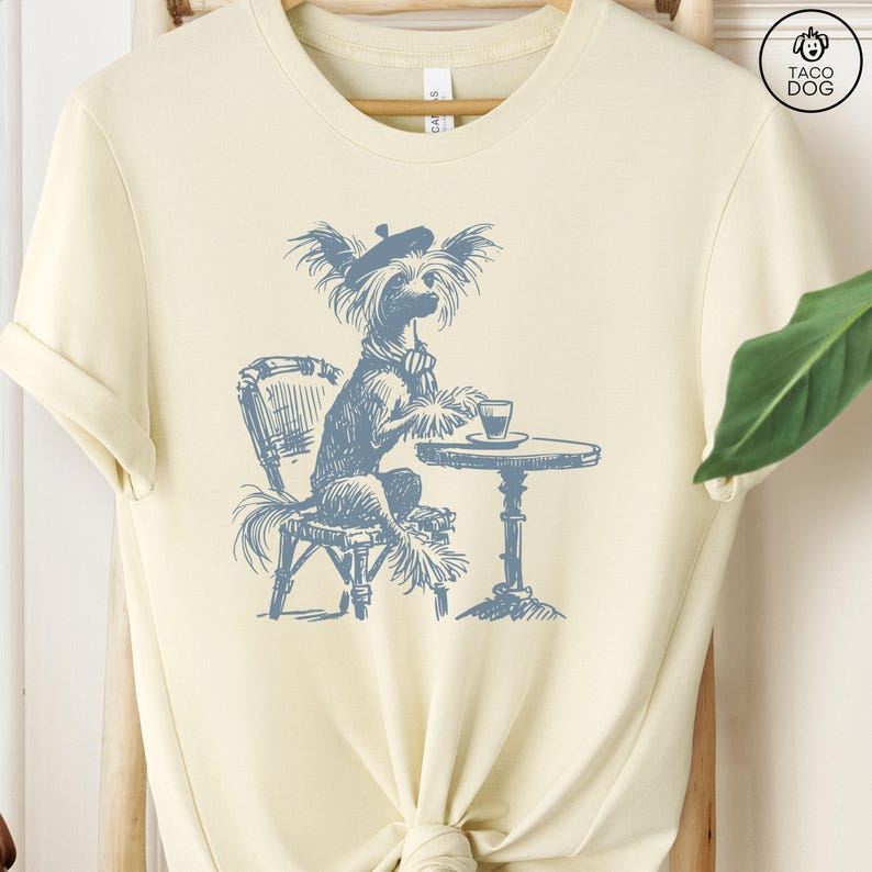 May include: A light yellow t-shirt with a blue illustration of a dog wearing a beret sitting at a table with a cup of coffee. The dog is a Chinese Crested dog. The text 'Taco Dog' is in the upper right corner of the image.