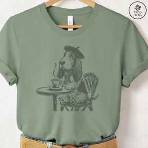 K&ouml;nnte beinhalten: Ein gr&uuml;nes T-Shirt mit einer Schwarzwei&szlig;illustration eines Basset Hounds, der einen Baskenm&uuml;tze tr&auml;gt und an einem Tisch mit einer Tasse Kaffee sitzt. Der Hund schaut nach links.