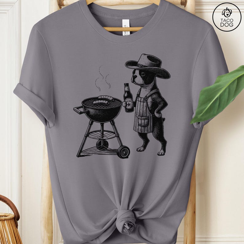 Peut inclure: Un t-shirt gris avec une illustration en noir et blanc d'un chien portant un chapeau de cowboy et un tablier, grillant des saucisses sur un barbecue au charbon de bois. Le chien tient une bouteille de bi&egrave;re.