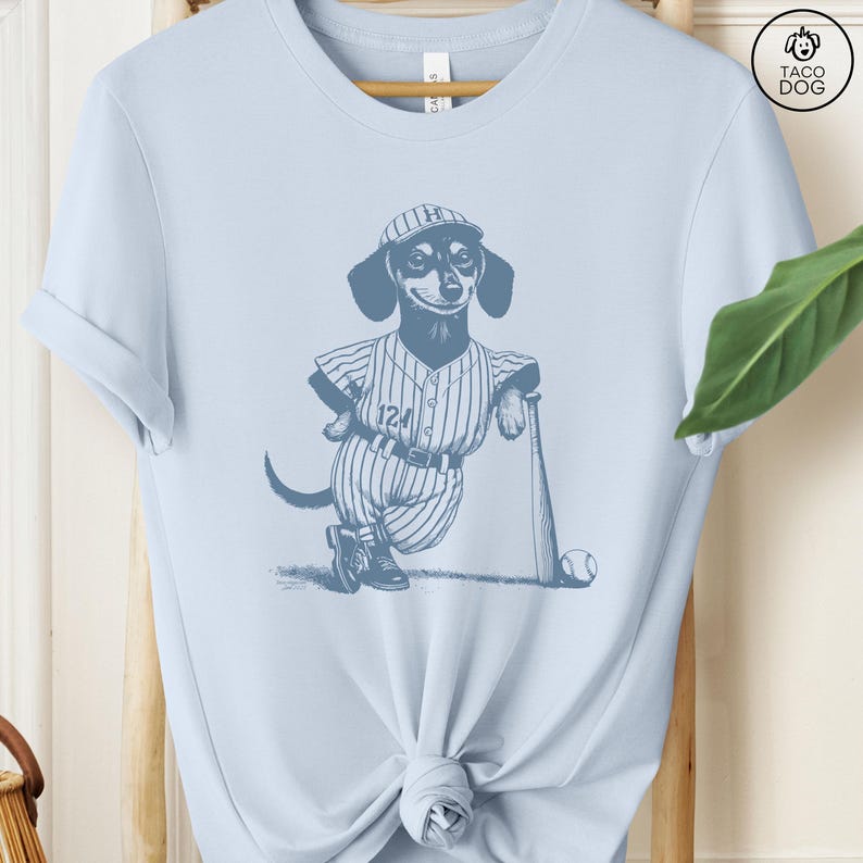 May include: A light blue t-shirt with a graphic of a dachshund wearing a baseball uniform and holding a bat. The dachshund is wearing a cap with the letter "H" on it and the number "124" on its jersey. The graphic is in shades of blue.
