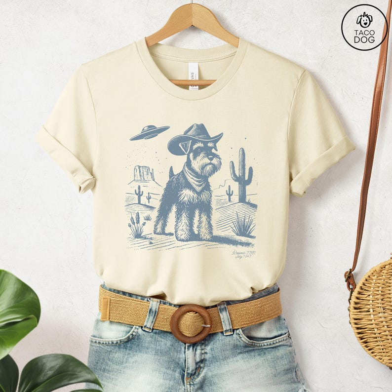 An off white t-shirt with a monochromatic blue illustration of a schnauzer wearing a cowboy hat standing in the desert with a UFO in the background.