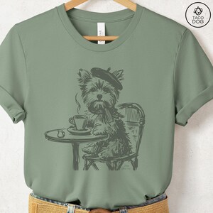 May include: Sage green t-shirt featuring a sketch of a dog wearing a beret and scarf, seated at a cafe table with a cup of coffee. The dog is sitting in a chair. The shirt has a crew neck and short sleeves. The brand logo is in the upper right corner.