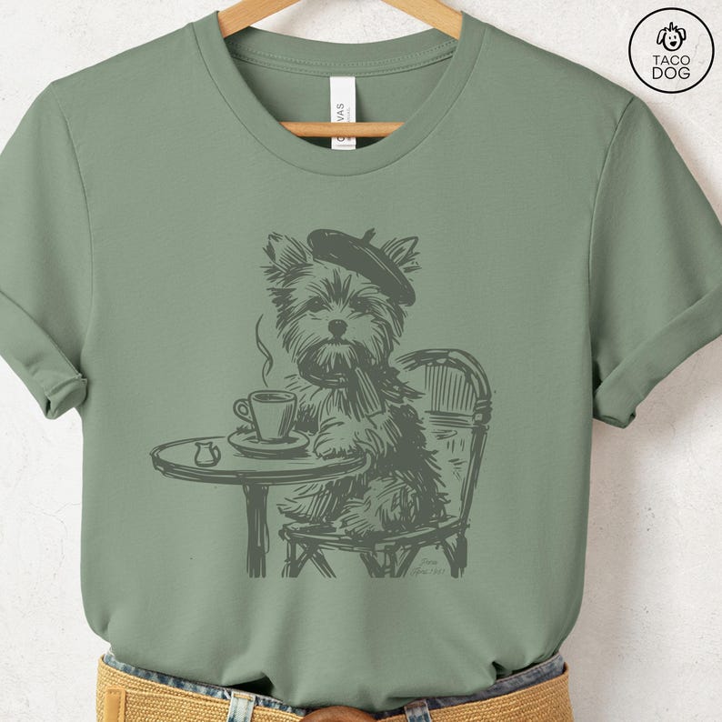 May include: Sage green t-shirt featuring a sketch of a dog wearing a beret and scarf, seated at a cafe table with a cup of coffee. The dog is sitting in a chair. The shirt has a crew neck and short sleeves. The brand logo is in the upper right corner.