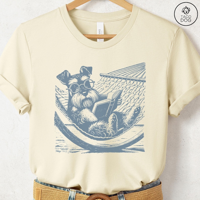 K&ouml;nnte beinhalten: Ein cremefarbenes T-Shirt mit einer blauen Illustration eines Hundes, der eine Brille tr&auml;gt und ein Buch liest, w&auml;hrend er in einer H&auml;ngematte entspannt.