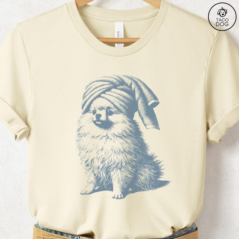 May include: Cream-colored t-shirt featuring a blue illustration of a Pomeranian dog wearing a towel on its head. The dog is sitting, with fluffy fur and a playful expression. The shirt is hanging on a wooden hanger.