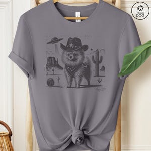 Puede incluir: Una camiseta gris con una ilustraci&oacute;n en blanco y negro de un perro con un sombrero de vaquero y un pa&ntilde;uelo. El perro est&aacute; de pie en un paisaje des&eacute;rtico con un cactus y un OVNI en el fondo.