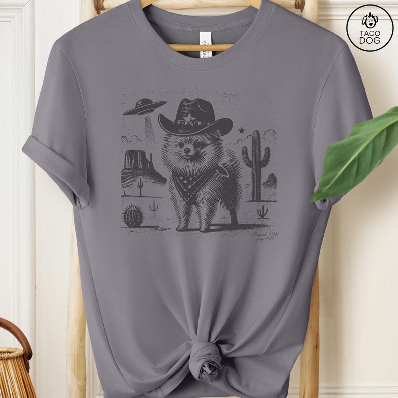 Puede incluir: Una camiseta gris con una ilustraci&oacute;n en blanco y negro de un perro con un sombrero de vaquero y un pa&ntilde;uelo. El perro est&aacute; de pie en un paisaje des&eacute;rtico con un cactus y un OVNI en el fondo.