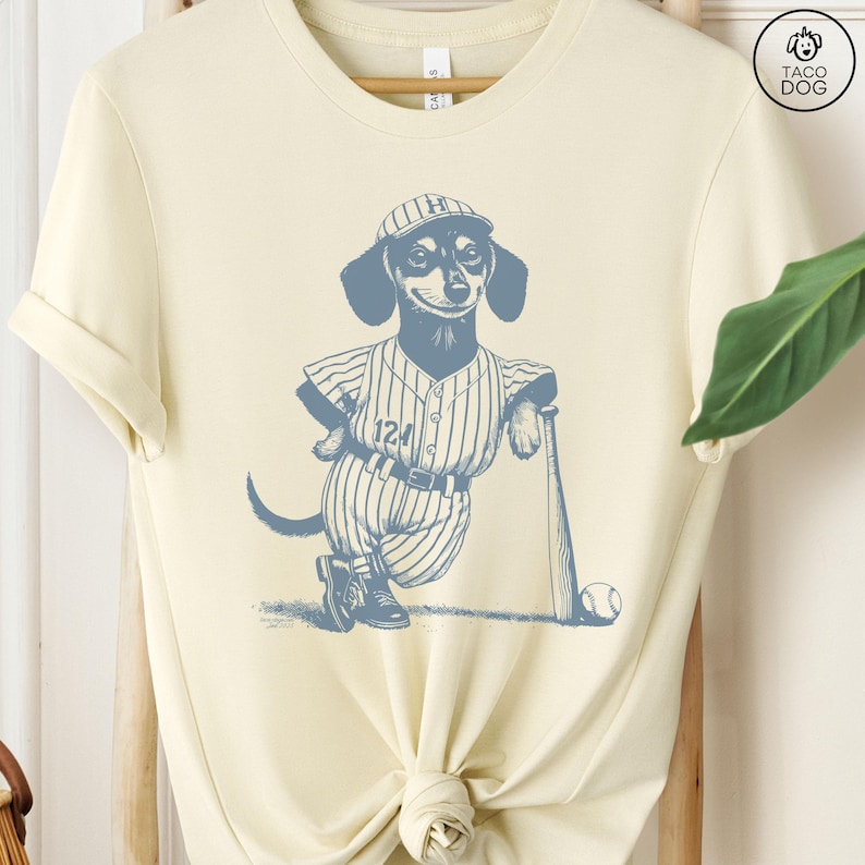 May include: A cream-colored t-shirt with a blue illustration of a dachshund wearing a baseball uniform and holding a bat. The dachshund is wearing a hat with the letter "H" on it and the number "124" on its jersey.