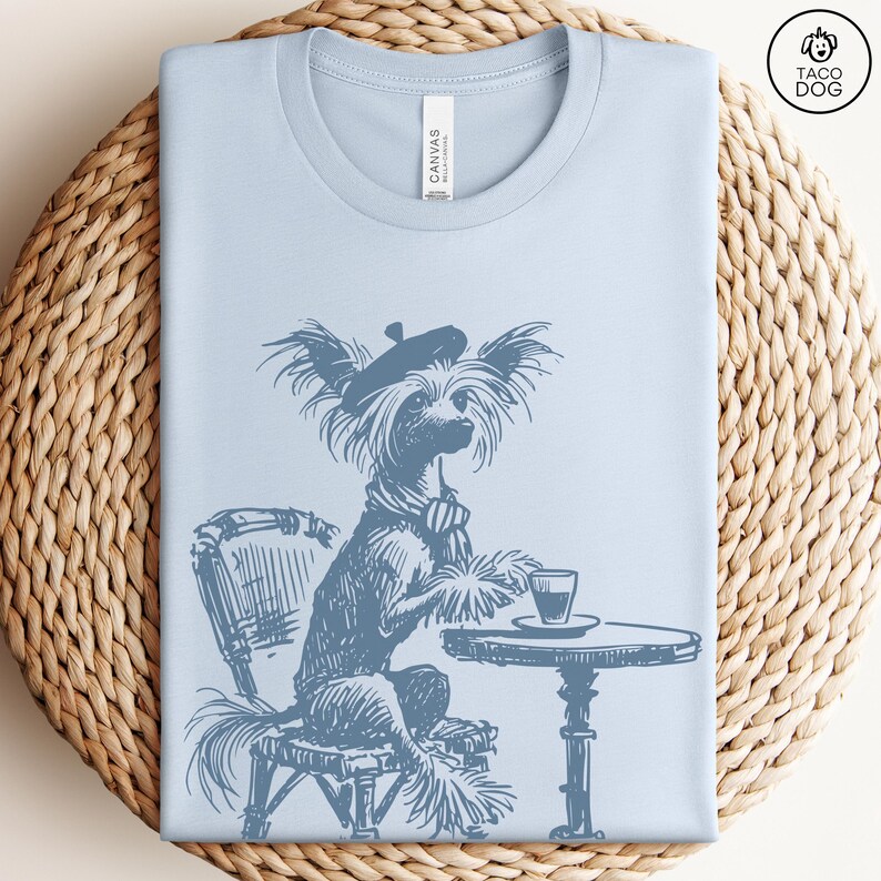 May include: A light blue t-shirt with a graphic of a dog wearing a beret and sitting at a table with a cup of coffee. The dog is a Chinese Crested.