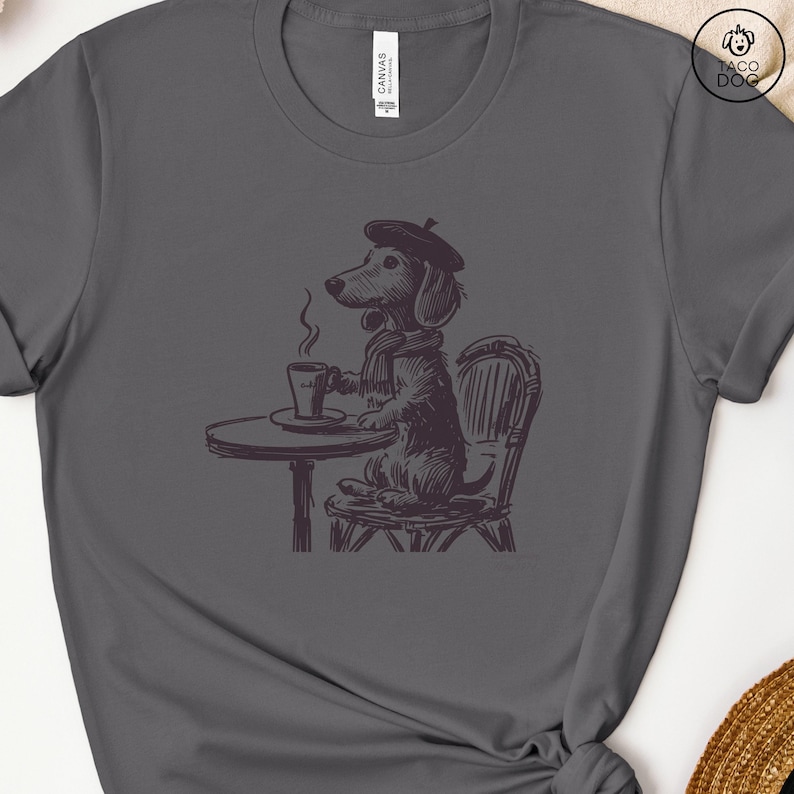 Może przedstawiać: Szary T-shirt z czarno-białą ilustracją  dacksa w berecie i szaliku, siedzącego przy stole z filiżanką kawy.