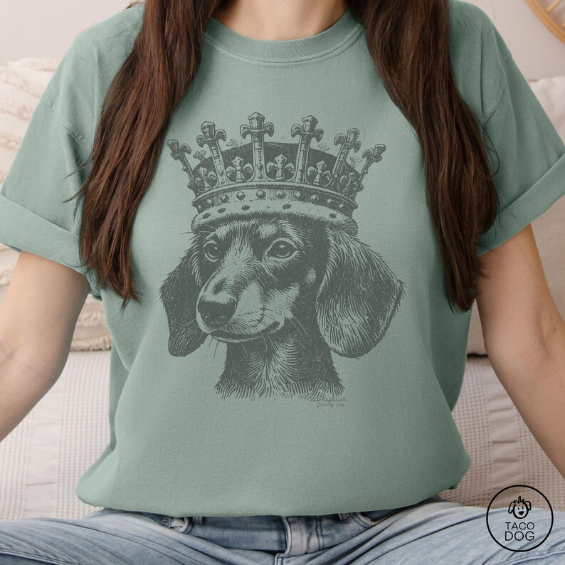 May include: A light green t-shirt with a black and white illustration of a dachshund wearing a crown. The crown has a detailed design with points and jewels.