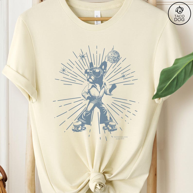 May include: A light yellow t-shirt with a blue graphic of a dog wearing roller skates and holding a disco ball. The dog is wearing sunglasses and a disco outfit. The graphic is surrounded by a sunburst design.