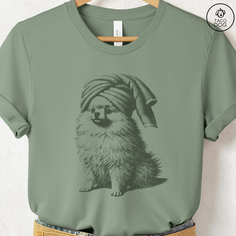 May include: Sage green t-shirt featuring a detailed illustration of a Pomeranian dog wearing a towel turban. The dog is depicted in a seated position, with the towel draped over its head. The shirt has a crew neck and short sleeves. The Taco Dog logo is in the upper right corner.
