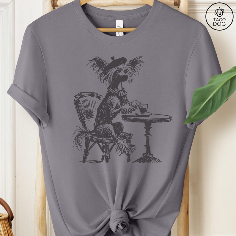 May include: A gray t-shirt with a black and white illustration of a dog wearing a beret and sitting at a table with a cup of coffee. The dog is a Chinese Crested.