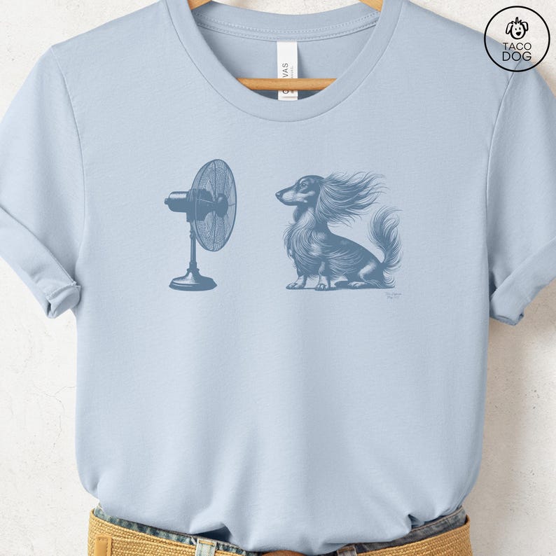 May include: Light blue t-shirt featuring a detailed illustration of a long-haired dachshund with wind-blown fur, facing a standing fan. The design is in a muted blue-grey color. The shirt has a crew neck and short sleeves. The brand logo "TACO DOG" is in the upper right corner.