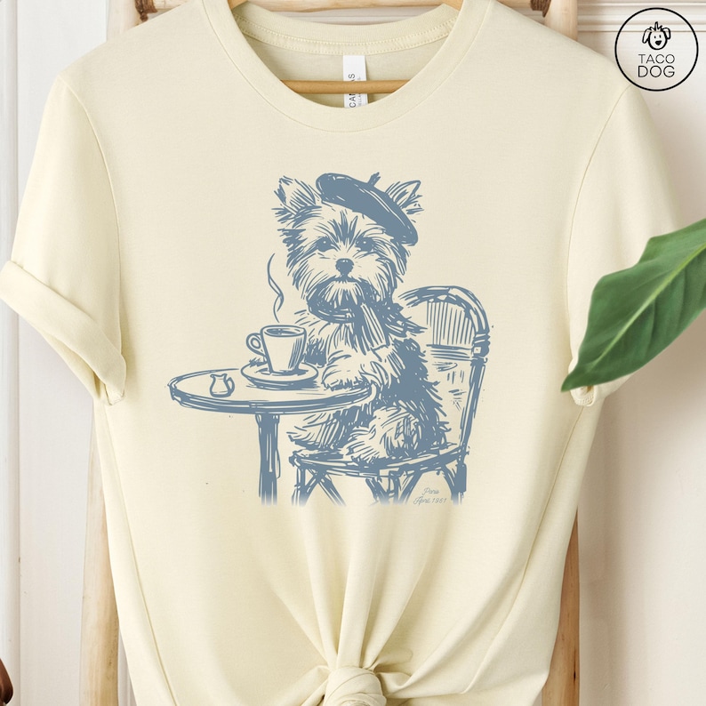 May include: A light yellow t-shirt with a blue illustration of a dog wearing a beret and sitting at a table with a cup of coffee. The text 'Taco Dog' is in the top right corner of the image. The text 'June 2023' is at the bottom of the image.