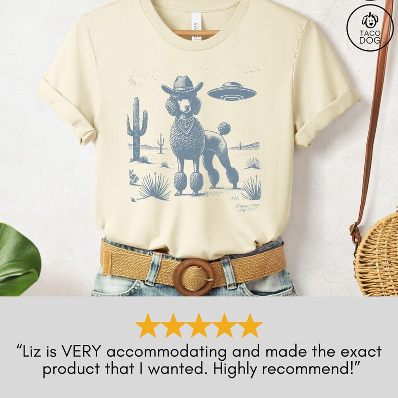 May include: Cream-colored t-shirt featuring a blue graphic of a poodle wearing a cowboy hat and a UFO. The design includes desert plants and a landscape. The shirt is on a hanger.